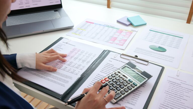 An individual at a desk reads paperwork while typing on a calculator.
